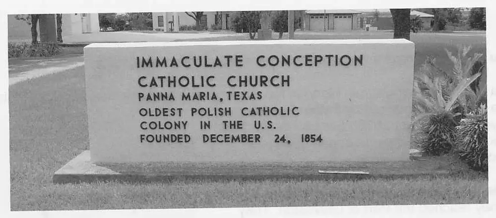 Photograph of a stone monument sign on a lawn reading 'Immaculate Conception Catholic Church, Panna Maria, Texas, Oldest Polish Catholic Colony in the U.S., Founded December 24, 1854', with buildings and trees in the background.