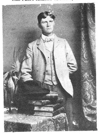 Studio portrait of a young man in a suit with bow tie, standing with hat and books on a table beside him.