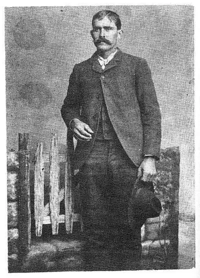 Studio portrait of a mustached man in a suit, standing beside a decorative chair, holding a hat.