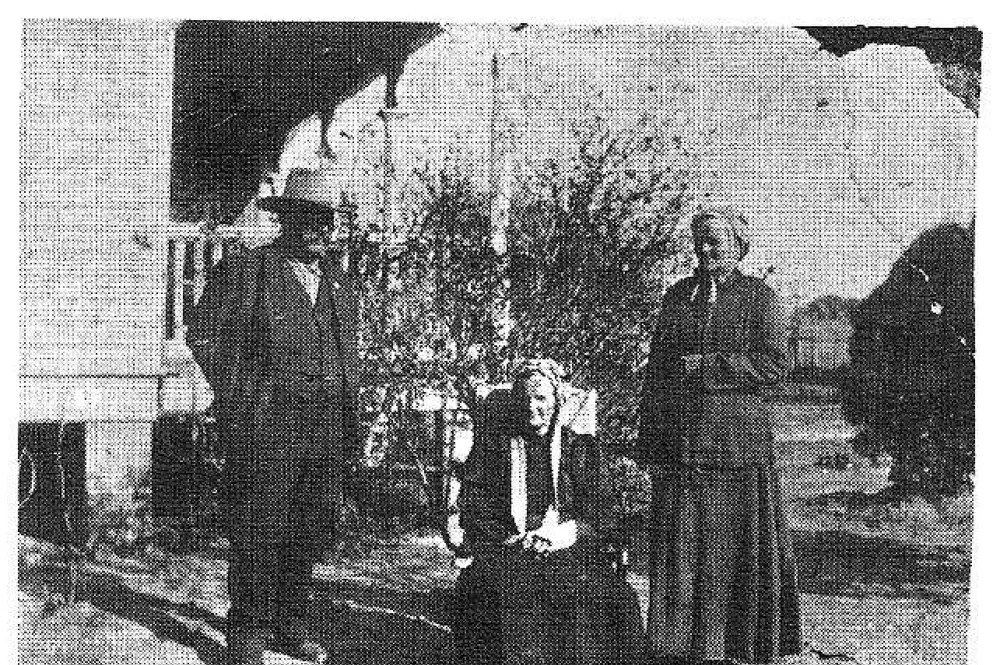 A man in a hat and suit and an elderly woman standing outdoors near a porch, with a smaller figure seated between them.