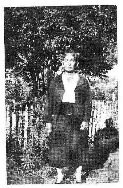 Woman standing outdoors in a dark coat and skirt in front of trees and a picket fence.