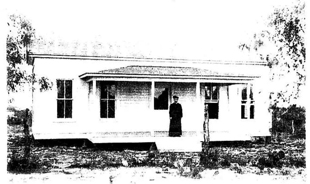 Black-and-white photograph of a small wood-frame house with a covered porch, a priest standing on the porch steps, trees around the property.