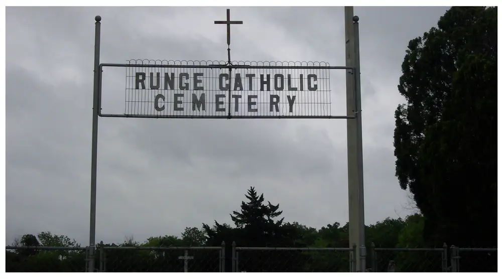 Cemetery entrance sign reading 'Runge Catholic Cemetery' with a cross above it, mounted between two posts under a cloudy sky.