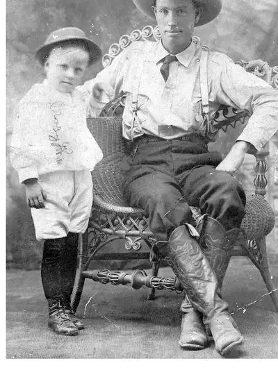 A young boy stands beside a man wearing a wide-brimmed cowboy hat, suspenders, and tall cowboy boots, seated in a wicker chair.