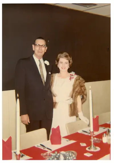 A man in a dark suit with a white boutonniere and a woman in a light dress with a fur stole, gloves, and a corsage, standing together at a banquet table set with red linens and lit candles.
