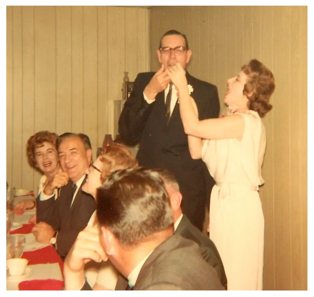 A man in glasses and suit feeding cake or food to a woman in a light dress at the head of the banquet table, with other guests seated nearby laughing.