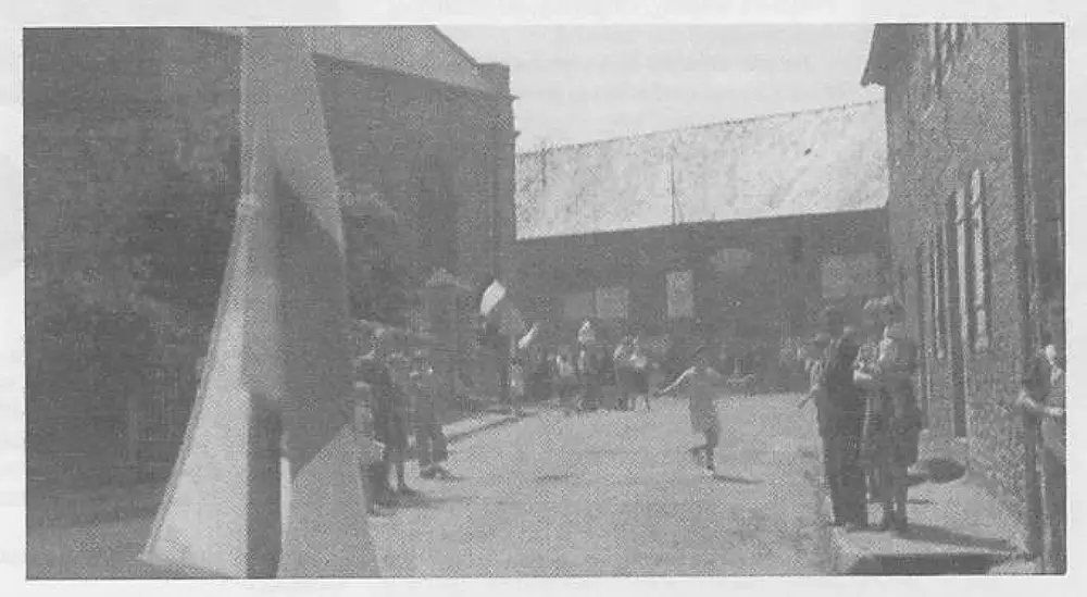 Street scene in a liberated French village in Brittany; townspeople lining the street to watch American troops pass, with a flag flying from a jeep in the foreground.