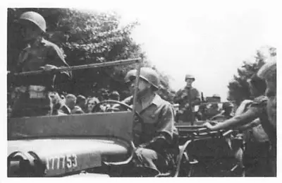 Soldiers in a military vehicle greeting a crowd of civilians lining the street, one soldier wearing a helmet standing in the vehicle.