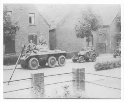 Armored car and jeep of the 15th Cavalry Squadron passing through a checkpoint with concrete anti-tank posts in Maasniel, in front of brick houses.