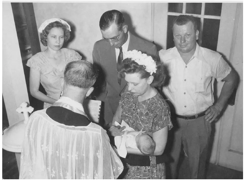 A priest in vestments, seen from behind, performing a christening over a baby held by its mother, surrounded by the godfather, godmother, and other family members standing indoors near a doorway.