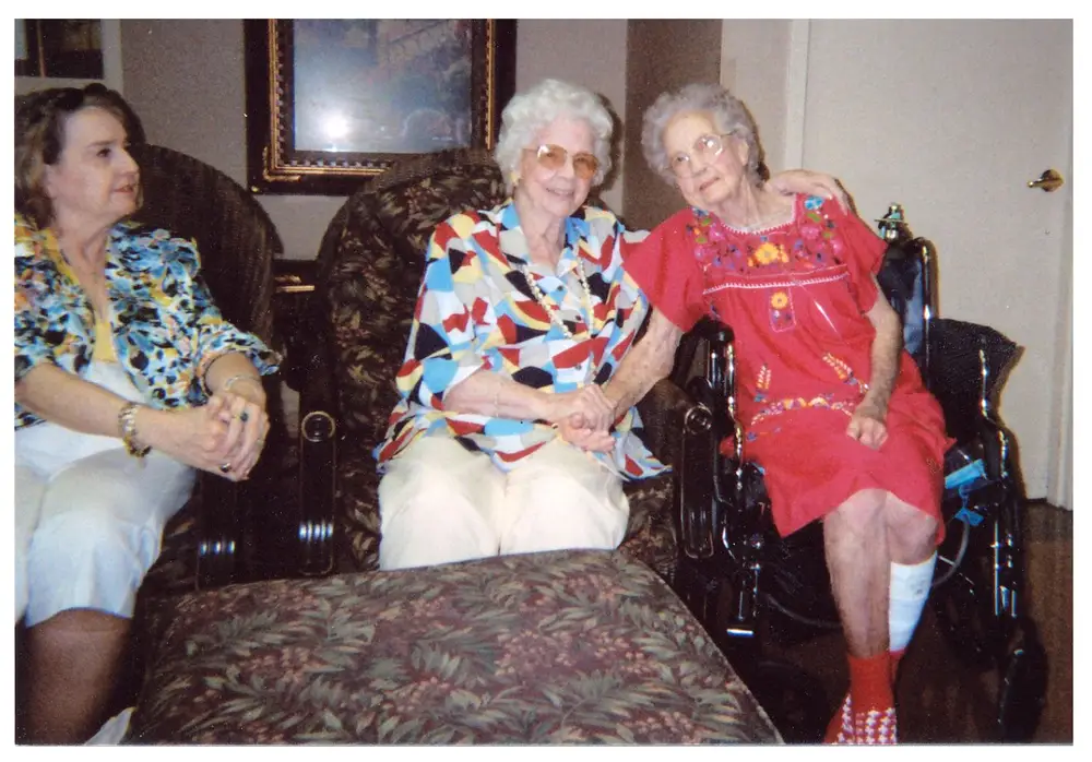 Three women seated in a living room; a younger woman sits at left, and two elderly women sit together in the center/right, one in a wheelchair with a bandaged leg.