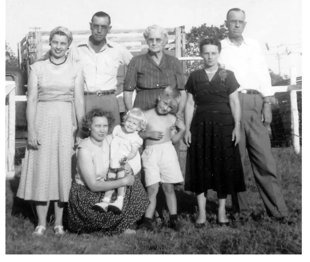 Black-and-white group photo of eight people (four men and women standing, an older woman, and two women seated with a young child and a small boy) posed outdoors in front of a wooden farm fence, from the 1952 Riley family get-together.