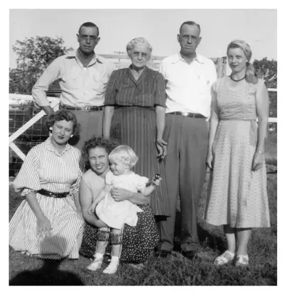 Black-and-white group photo of seven people (three men standing, an older woman, and two women, one holding a toddler) posed outdoors in front of a wooden farm fence, from the 1952 Riley family get-together.