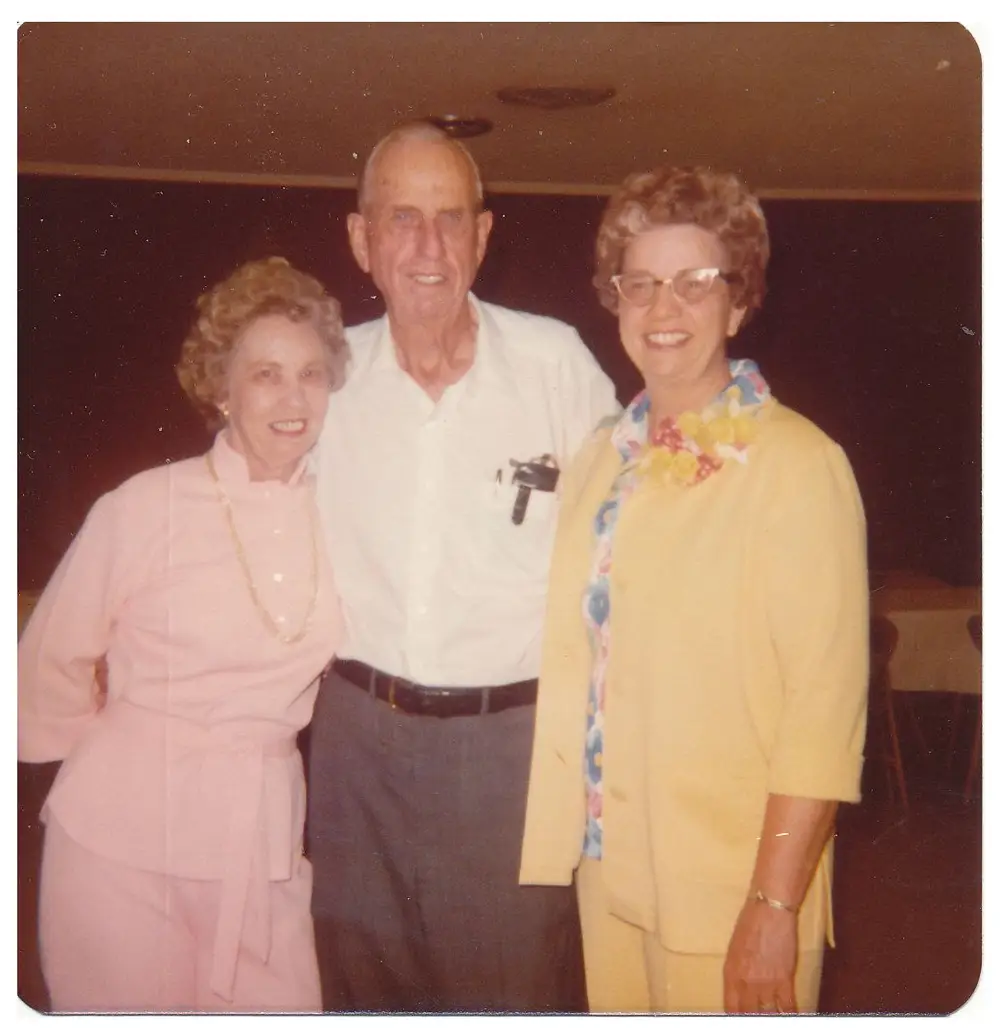 Color photo of three older adults (a woman in pink, a man in a white shirt, and a woman in a yellow floral-trimmed cardigan) standing together indoors, smiling for the camera, from the 1970's.