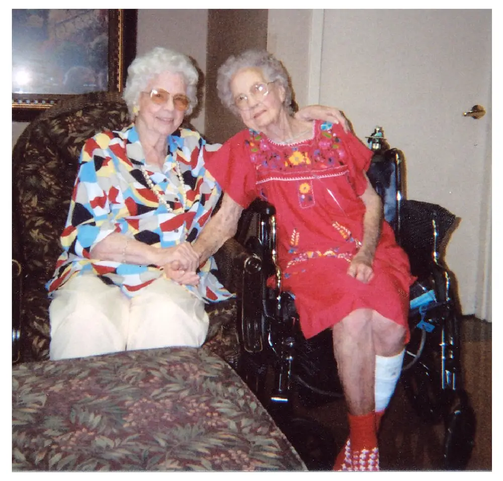 Two elderly women seated together holding hands, one in a colorful geometric-print blouse and white pants, the other in a red embroidered dress with a bandaged leg