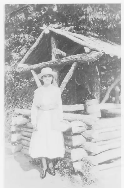 A young woman in a white hat and long white dress stands beside a rustic log-built wishing well with a bucket, under trees.